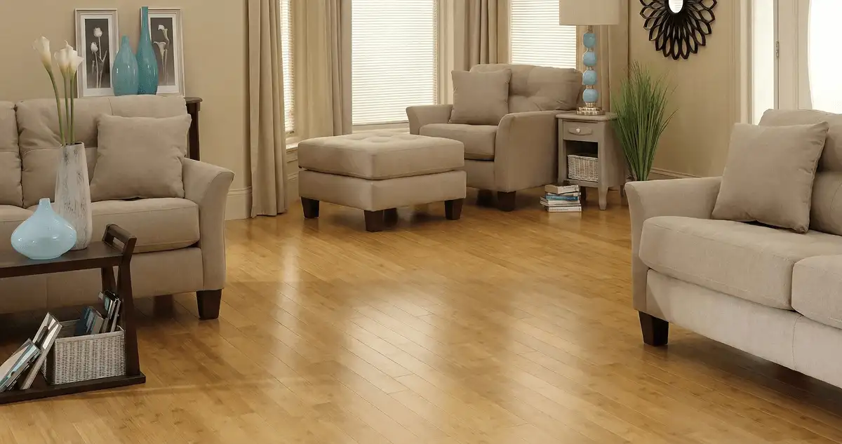 A living room with professionally installed bamboo flooring, beige sofas, a matching ottoman, a side table with a blue lamp, vases, framed pictures, and large windows with blinds and curtains in Columbus, GA